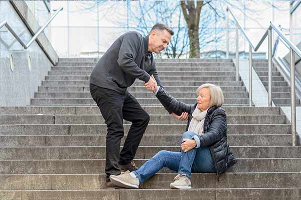 A man helps an elderly woman seated on a concrete staircase. She holds his hand as he assists her in standing. They are outdoors, and the background shows a tree and railing beside the stairs.