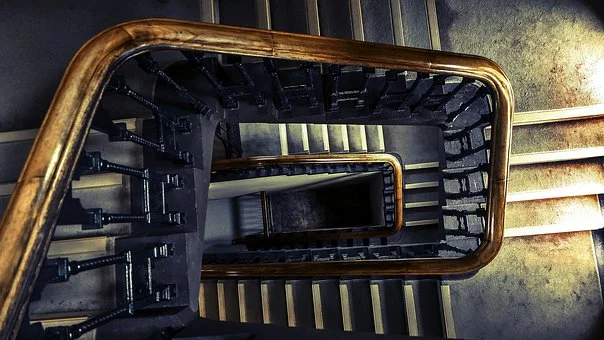 A spiraling stairwell viewed from above showcases multiple floors with a wooden handrail and dark balusters. Stone steps create dramatic shadows and highlights, enhancing the structure's architectural beauty.