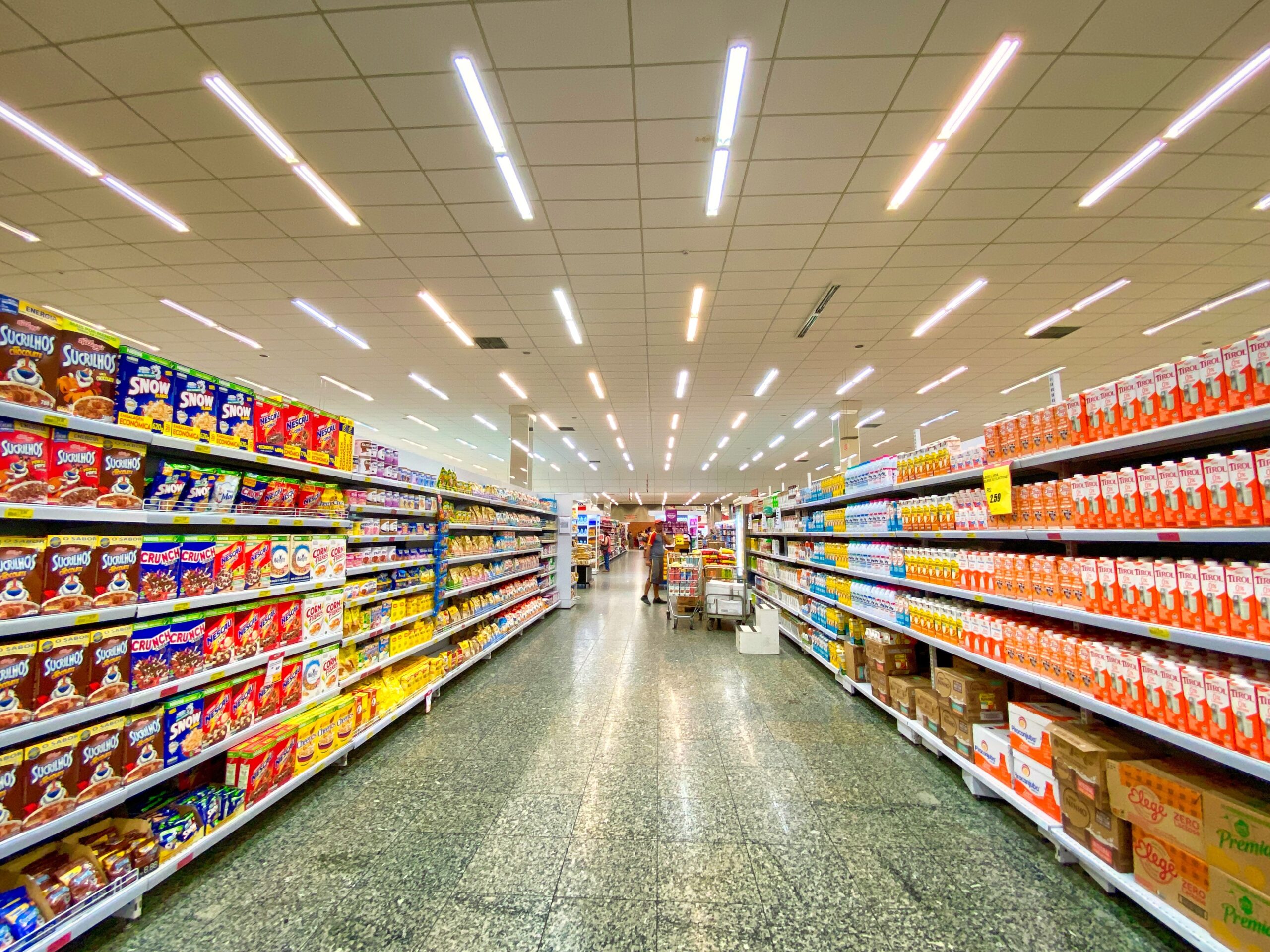 A wide-angle view of a supermarket aisle reveals well-organized shelves. The left displays various cereal boxes, while the right side showcases stacked cartons of juice. Bright overhead lights illuminate the aisle, which stretches into the distance—reassurance for those seeking clarity after a supermarket accident.