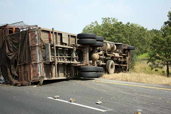 An overturned truck lies on its side on the road shoulder. The truck's wheels are visible, and there is a small amount of debris scattered on the roadway. Trees and vegetation are seen in the background under a clear sky.