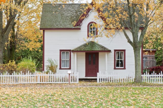A charming white house with red trim sits behind a white picket fence in a quaint New Jersey neighborhood. The front features two windows and a red door with a small porch. Autumn leaves cover the ground, and two trees with fall foliage frame the scene, offering an ideal Airbnb escape.