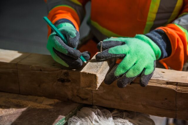 Amidst a bustling workshop vibrant with sawdust, a person in bright green gloves and an orange safety jacket carefully marks a piece of wood with pencil precision. It's crucial to stay focused in such environments, where the guidance of work injury attorneys can be essential if accidents occur.