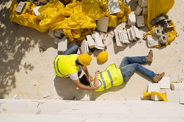 Two construction workers are on the ground. One worker is lying down surrounded by bricks and yellow bags, while the other is kneeling beside him. Both are wearing yellow safety vests and helmets. Shadows of trees are visible.