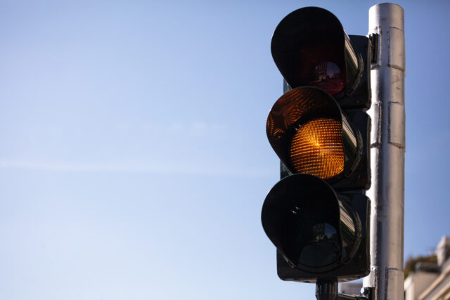 A traffic light set against a clear blue sky displays a yellow light, indicating caution. The metal structure holds three circular lamps, with the yellow light illuminated in the center—a reminder of how faulty traffic signals can prompt extra vigilance.