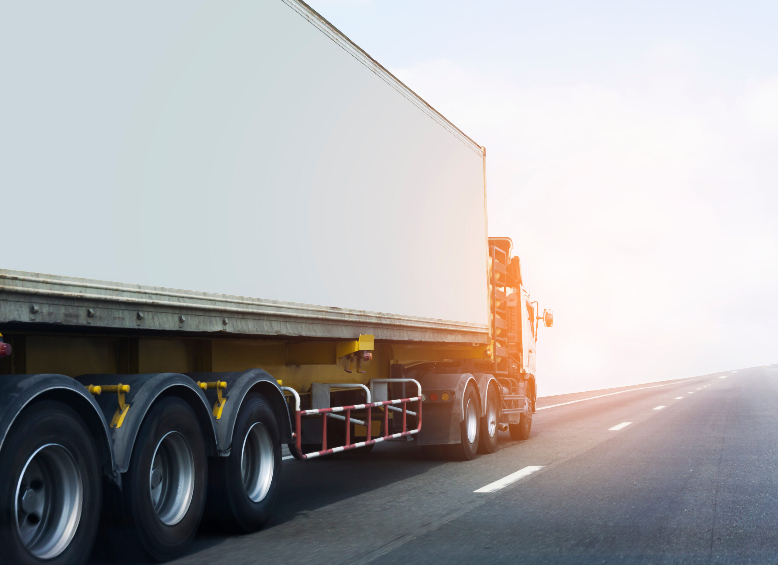 A large semi-truck with a white trailer cruises along an empty highway under a clear sky. The sun shines brightly, casting light on the road and vehicle, emphasizing the scene's sense of motion and travel.