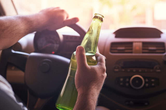 A person grips the steering wheel of a car with their left hand, while a green glass bottle rests in their right. The dashboard, featuring the speedometer and radio, is visible—a stark reminder of the dangers posed by drunk drivers in NJ.