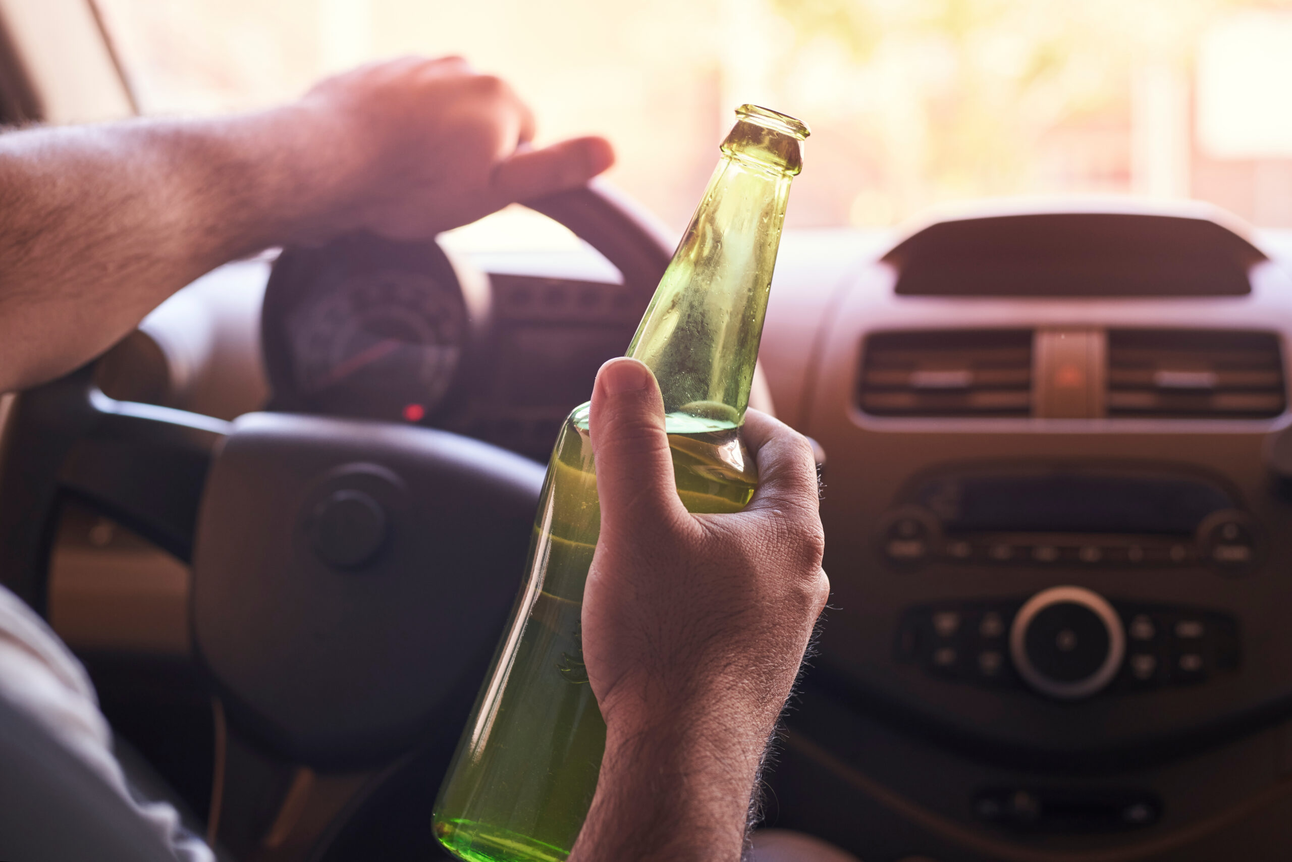 A person grips the steering wheel of a car with their left hand, while a green glass bottle rests in their right. The dashboard, featuring the speedometer and radio, is visible—a stark reminder of the dangers posed by drunk drivers in NJ.