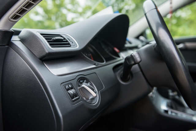 Close-up of a car interior showing the steering wheel, headlight control dial, and air vent. The dashboard is partially visible with devices recording black box data, while the view outside is blurred with green foliage.