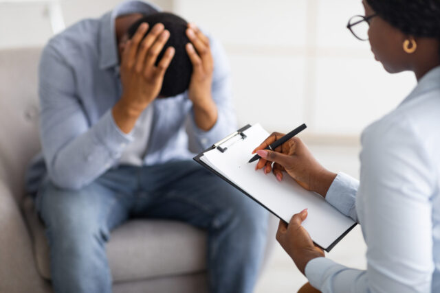A person wearing a blue shirt is seated, holding their head in their hands, overwhelmed by memories of a car accident. Across from them, another person in a light-colored shirt holds a clipboard and pen, appearing to take notes in the counseling session focused on managing PTSD symptoms.